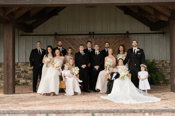 A professional wedding party portrait featuring the bride, groom, and their wedding party posing in front of a rustic wooden barn structure.