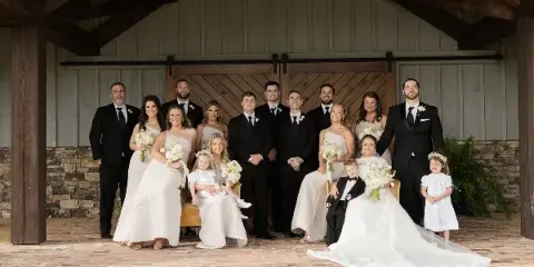 A professional wedding party portrait featuring the bride, groom, and their wedding party posing in front of a rustic wooden barn structure.