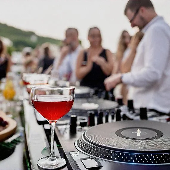A DJ in a white shirt works a turntable setup at an outdoor event with a glass of red drink in the foreground and guests in the background.