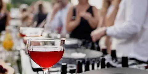 A DJ in a white shirt works a turntable setup at an outdoor event with a glass of red drink in the foreground and guests in the background.