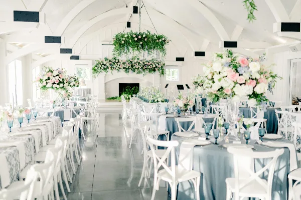 White modern farmhouse wedding reception room with long tables, white chairs, floral centerpieces, hanging floral installations, and a fireplace at the far end.