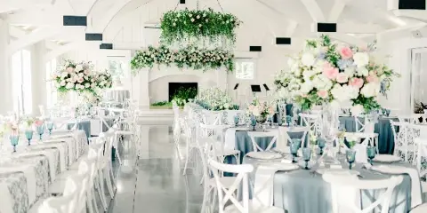White modern farmhouse wedding reception room with long tables, white chairs, floral centerpieces, hanging floral installations, and a fireplace at the far end.