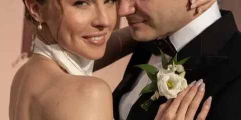 A bride with an elegant updo hairstyle and full makeup embracing a groom in a tuxedo.