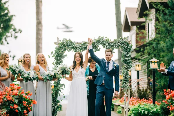 A bride and groom holding hands and raising their arms in celebration under a floral arch, surrounded by bridesmaids and groomsmen.