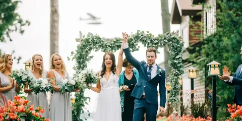 A bride and groom holding hands and raising their arms in celebration under a floral arch, surrounded by bridesmaids and groomsmen.