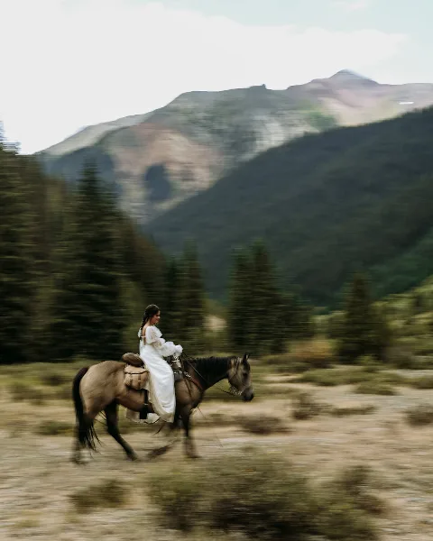 A bride in a white dress riding a horse in a mountainous landscape with evergreen trees.