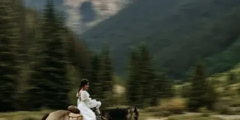 A bride in a white dress riding a horse in a mountainous landscape with evergreen trees.