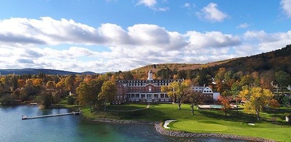 Aerial view of The Otesaga Resort Hotel on the shores of Otsego Lake with autumn foliage and a blue sky.