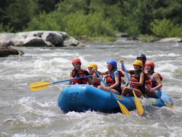 A group of people in helmets and life jackets paddling a blue raft through white water rapids on a river.