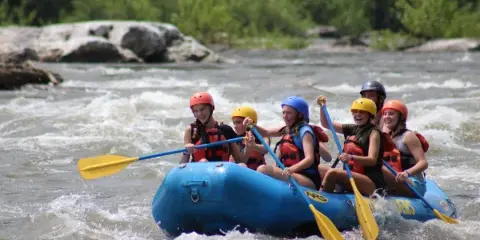 A group of people in helmets and life jackets paddling a blue raft through white water rapids on a river.