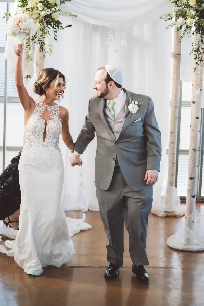 A bride in a white lace gown and a groom in a grey suit holding hands and smiling in a decorated wedding venue.
