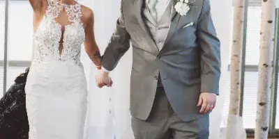 A bride in a white lace gown and a groom in a grey suit holding hands and smiling in a decorated wedding venue.