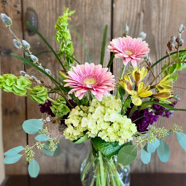 A mixed floral arrangement featuring pink gerbera daisies, yellow alstroemeria, and green hydrangeas in a clear glass vase.