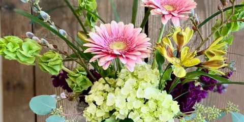 A mixed floral arrangement featuring pink gerbera daisies, yellow alstroemeria, and green hydrangeas in a clear glass vase.