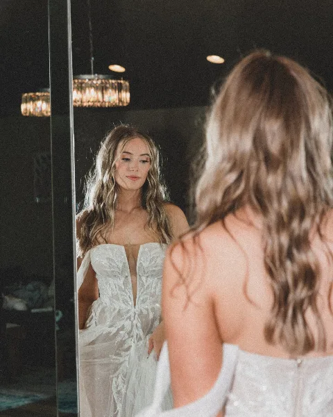 A bride in a white wedding dress looking at her reflection in a mirror.