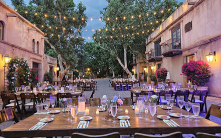 An outdoor courtyard venue set up for a wedding reception with long wooden tables, floral centerpieces, and string lights.