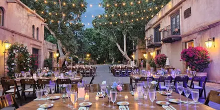 An outdoor courtyard venue set up for a wedding reception with long wooden tables, floral centerpieces, and string lights.