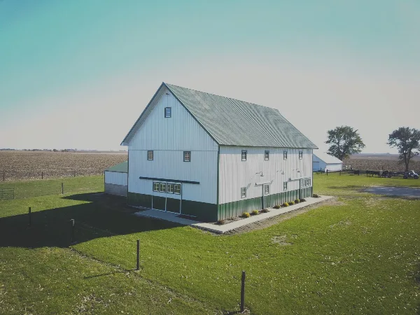 A white barn with a green metal roof and green trim sits on a grassy lawn surrounded by open fields.