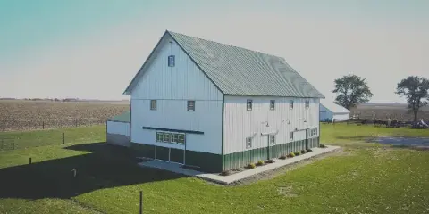 A white barn with a green metal roof and green trim sits on a grassy lawn surrounded by open fields.