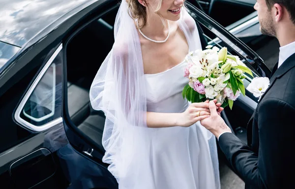 A bride and groom standing next to a black luxury car, with the bride holding a bouquet.
