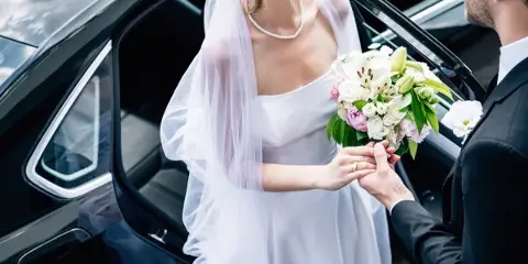 A bride and groom standing next to a black luxury car, with the bride holding a bouquet.