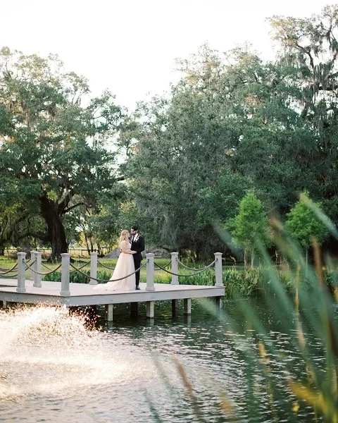 A bride and groom standing on a wooden dock by a pond surrounded by trees and greenery.