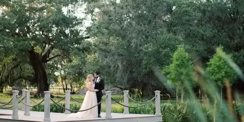 A bride and groom standing on a wooden dock by a pond surrounded by trees and greenery.
