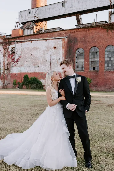 A bride and groom standing together in a field in front of a brick building.