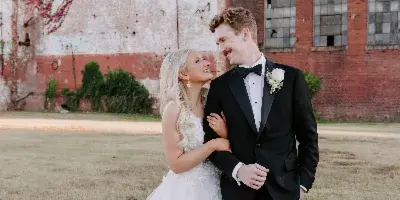A bride and groom standing together in a field in front of a brick building.