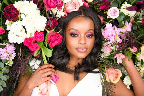 A professional portrait of a woman with dark hair and pink makeup, framed by a large, colorful floral arrangement of roses and hydrangeas.