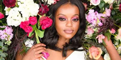 A professional portrait of a woman with dark hair and pink makeup, framed by a large, colorful floral arrangement of roses and hydrangeas.