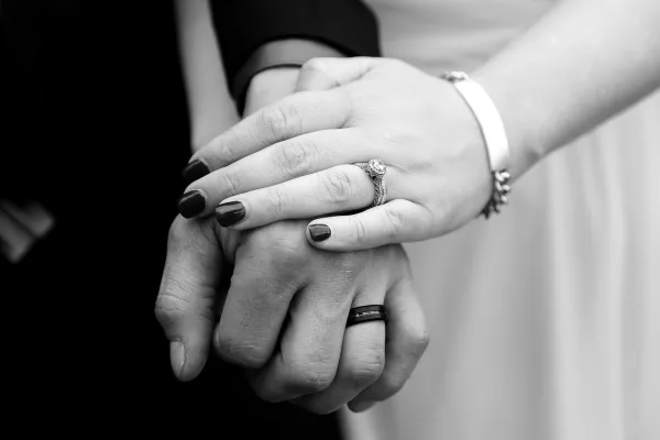Black and white close-up of a couple's hands holding each other, showing an engagement ring and a wedding band.