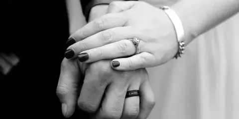 Black and white close-up of a couple's hands holding each other, showing an engagement ring and a wedding band.