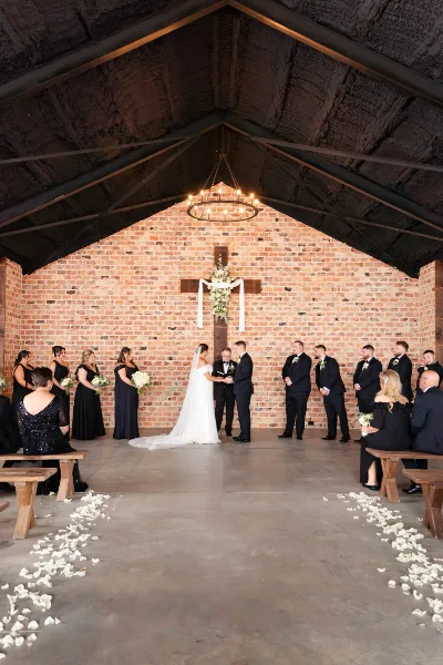 A wedding ceremony in progress at an open-air chapel with a brick wall backdrop, wooden cross, and chandelier lighting.