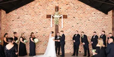 A wedding ceremony in progress at an open-air chapel with a brick wall backdrop, wooden cross, and chandelier lighting.