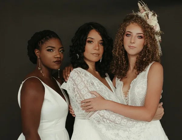 Three women in white dresses posing together, showcasing professional bridal hair and makeup styles.