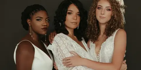 Three women in white dresses posing together, showcasing professional bridal hair and makeup styles.