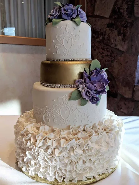A three-tiered wedding cake featuring white ruffles, a gold middle tier, and purple floral decorations.