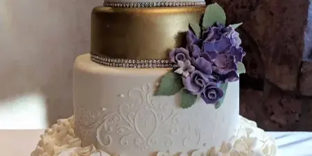 A three-tiered wedding cake featuring white ruffles, a gold middle tier, and purple floral decorations.