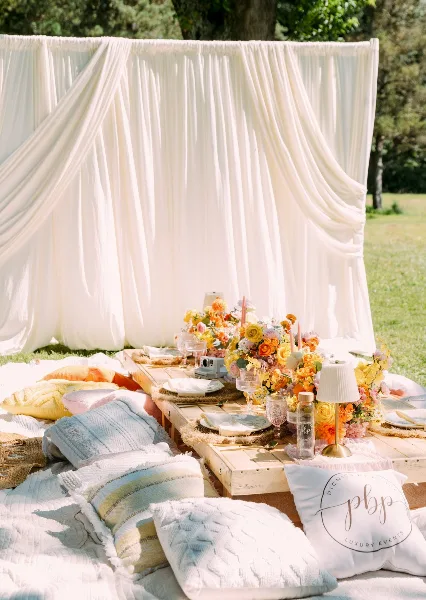 Outdoor event setup featuring white draping, a floral centerpiece, and styled pillows on a wooden table.