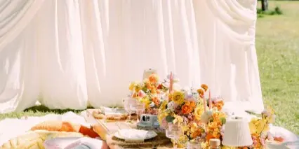 Outdoor event setup featuring white draping, a floral centerpiece, and styled pillows on a wooden table.