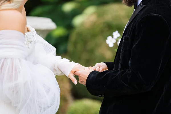 A close-up photograph of a bride and groom holding hands and exchanging rings during a wedding ceremony.