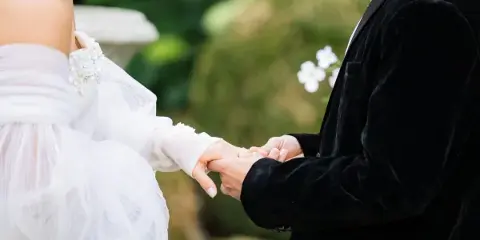 A close-up photograph of a bride and groom holding hands and exchanging rings during a wedding ceremony.