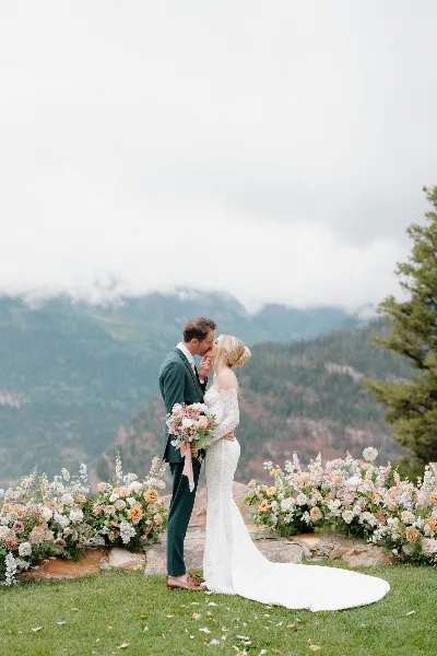 A bride and groom kissing in front of a mountain backdrop with floral arrangements.