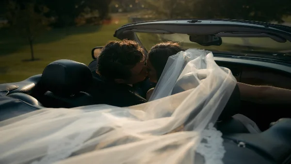 Cinematic still of a bride and groom kissing in a convertible car with the bride's veil flowing.