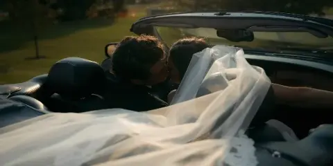 Cinematic still of a bride and groom kissing in a convertible car with the bride's veil flowing.