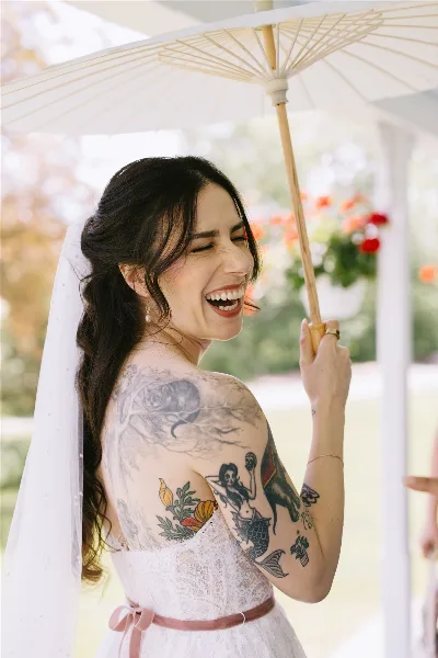 A smiling bride in a white lace dress and veil holding a paper parasol, with styled hair and makeup.