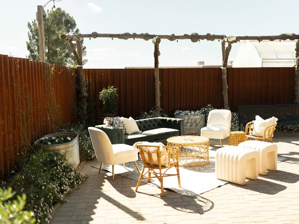 Rustic outdoor patio area featuring a wooden pergola, green sofa, white chairs, and potted plants.