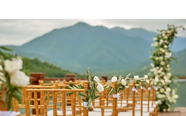 A wedding ceremony setup featuring bamboo chairs decorated with white flowers and greenery, set against a scenic mountain background.