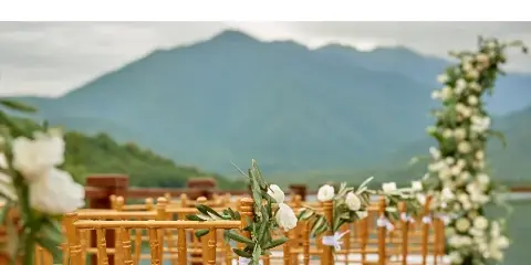 A wedding ceremony setup featuring bamboo chairs decorated with white flowers and greenery, set against a scenic mountain background.